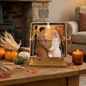 May include: A gold-toned metal rotating picture frame featuring a photograph of two people standing in an autumn forest. The frame is mounted on a rectangular base and includes side knobs for rotation, displaying a warm, seasonal aesthetic.