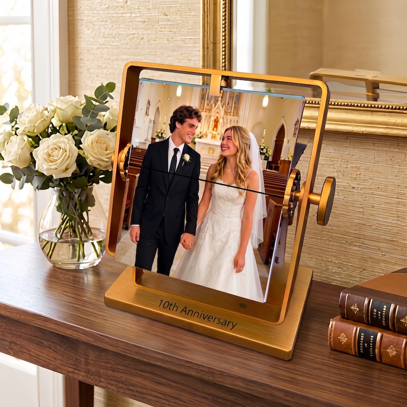 May include: A gold-toned metal rotating picture frame with a square base engraved with the text 10th Anniversary. The frame holds a photograph of a person in a black suit and a person in a white wedding dress standing in a church.