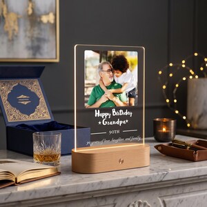 May include: A rectangular acrylic plaque with an illuminated edge, set in a light wood base. The plaque features a printed photograph of an older person and a child, with the text "Happy Birthday Grandpa 90TH 90 years of love, laughter and family."