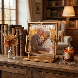 A luxury gold rotating picture frame on a vintage wooden desk with a lit candle and old books, displaying an elderly couple hugging for a meaningful 50th golden wedding anniversary gift.