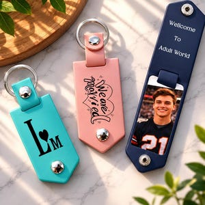 May include: Three rectangular keychains with metal rings and snap buttons. One is turquoise with the letters LM and a heart, one is pink with the text We are married, and one is navy blue with the text Wellcome To Adult World and a photo of a person.