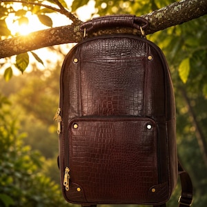 Könnte beinhalten: Ein dunkelbrauner Rucksack mit einer strukturierten Krokodilleder-Optik. Der Rucksack verfügt über einen Tragegriff, ein großes Hauptfach, eine kleinere Vordertasche sowie goldfarbene Metallbeschläge an den Ecken und Reißverschlüssen.