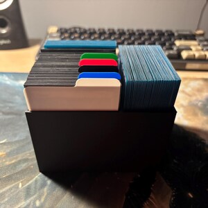 May include: A black card organizer with multiple stacks of cards. The cards are separated by colored dividers: white, blue, red, green, and black. The cards are blue and black. A keyboard is in the background.