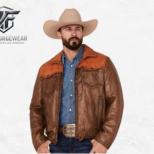 Op de afbeelding: Bruine leren jas met een bruinbeige pas en kraag, gedragen met een spijkeroverhemd en cowboyhoed. De jas heeft twee voorzakken en een decoratieve gesp. Het Nightforge Wear-logo is zichtbaar.