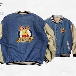 Könnte beinhalten: Zwei Jeans-Collegejacken mit beigefarbenen Ärmeln. Eine Jacke hat ein großes, gesticktes Winnie-the-Pooh-Design auf dem Rücken mit dem Text "Pooh" und "The Sort of Bear Who's Always There". Die andere Jacke hat ein kleineres Pooh-Design.