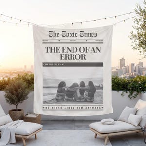 May include: A large, off-white tapestry with newspaper-style text. The headline reads "The Toxic Times" and "THE END OF AN ERROR." A black and white photo shows four people on a beach. The text "WE NEVER LIKED HIM ANYWAYS" is at the bottom.
