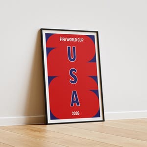 May include: A framed vertical poster featuring a red background with large, stylized navy blue numbers 2026 overlapping the text. The words FIFA WORLD CUP appear at the top in white, with the letters U S A stacked vertically in blue in the center.