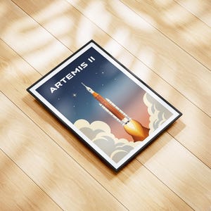 May include: Framed art print featuring a rocket launching into space, with the text "ARTEMIS II" at the top. The artwork has a retro style, with a color palette of orange, blue, and white. The print is in a black frame.