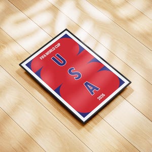 May include: A rectangular framed poster featuring a dark blue background with large, overlapping red circular shapes. The text FIFA WORLD CUP is printed in white at the top, with the letters USA stacked vertically in blue and the year 2026 in white.