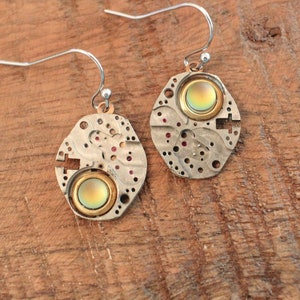 May include: A pair of unique earrings featuring a steampunk design. Each earring has a silver-toned, oval-shaped base made from repurposed watch parts. A circular, iridescent stone is set in the center of each earring, surrounded by a gold-toned metal ring.