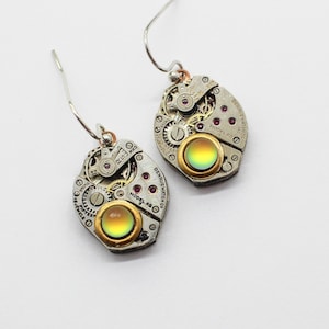 May include: A pair of steampunk-style earrings. Each earring features a watch mechanism with silver-tone metal, small red accents, and a gold-tone bezel surrounding a color-shifting, iridescent stone. The earrings hang from silver-tone ear wires.