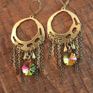 Puede incluir: Un par de pendientes de color latón con un diseño circular y cadenas colgantes. Cada pendiente presenta un colgante de cristal iridiscente grande. Los pendientes tienen un cierre de palanca y se exhiben sobre una superficie de madera.