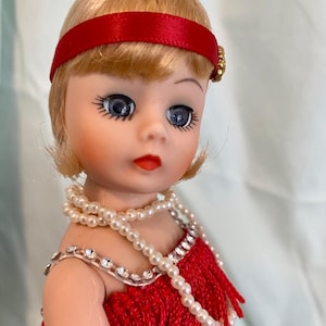 May include: A vintage doll with blonde hair, blue eyes, and red lips. The doll wears a red headband and a red fringed dress, adorned with a pearl necklace and rhinestone accents. The doll's features are detailed, and the overall aesthetic is reminiscent of a bygone era.