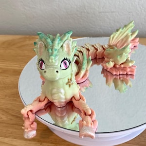 May include: A whimsical, pastel-colored dragon figurine with a pearlescent finish. The dragon features a light green head and tail, with pink and yellow accents. It is posed on a mirrored surface, showcasing its reflection.