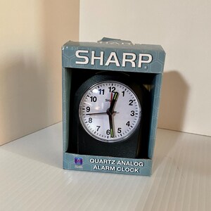 May include: A black Sharp quartz analog alarm clock inside its original blue and grey cardboard packaging. The clock face is white with black numerals, black hour and minute hands, and a red second hand. The box displays the text SHARP.