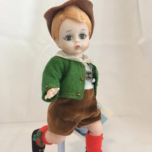 May include: A vintage doll dressed in traditional attire. The doll has red hair and is wearing a brown hat, green jacket, brown shorts, red socks, and black shoes. The jacket has gold buttons and a white collar. The doll is posed as if walking.