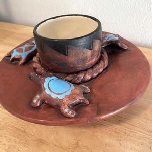 May include: A handcrafted, ceramic serving dish with a central cup and a wide, round base. The dish is primarily reddish-brown with a black rim on the cup. Decorative turtle figures with blue accents are placed around the rim. The dish is designed for serving.