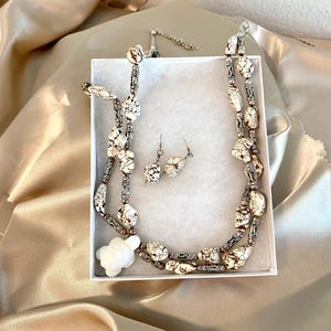 May include: A necklace and earring set displayed in a white box. The necklace features cream-colored, speckled beads with silver-toned accents. A white turtle pendant is at the bottom. The matching earrings have similar speckled beads.