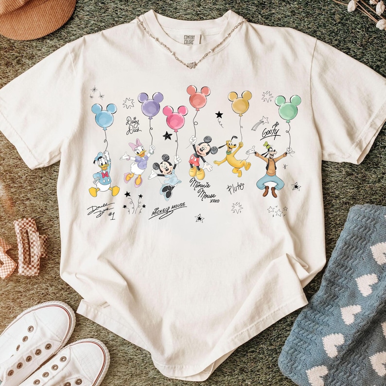 May include: Cream-coloured t-shirt with a vibrant graphic of Disney characters holding Mickey Mouse-shaped balloons. The design includes Donald Duck, Daisy Duck, Mickey Mouse, Minnie Mouse, Pluto, and Goofy. The shirt has a crew neck and short sleeves.