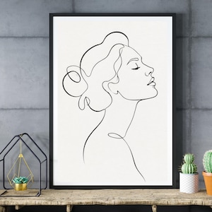 Könnte beinhalten: Gerahmter Kunstdruck mit einer minimalistischen Strichzeichnung des Profils einer Frau. Das Kunstwerk ist eine durchgehende Linie, die Gesicht, Hals und Haare in einem einfachen, eleganten Stil darstellt. Der Hintergrund ist weiß.