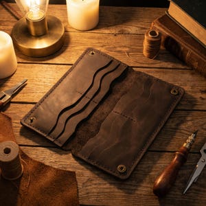 May include: Open brown leather wallet with multiple card slots and a snap closure. The wallet is displayed open, revealing the interior design. The leather has a rich, textured appearance, suggesting quality craftsmanship. The wallet is on a wooden surface.