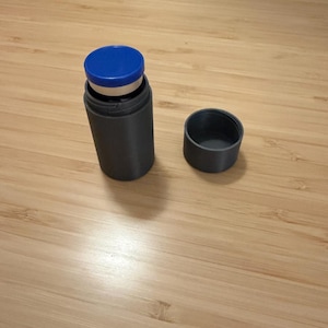 Op de afbeelding: Een cilindrische, donkergrijze container met een blauw deksel en een aparte dop. De container bevat een kleiner, witgekleurd object. De items zijn geplaatst op een lichtbruin houten oppervlak.