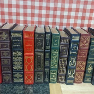 May include: A collection of antique books with decorative covers. The books are various colors, including red, green, and blue, with gold lettering and ornate designs. The books are arranged in a row against a red and white checkered background.