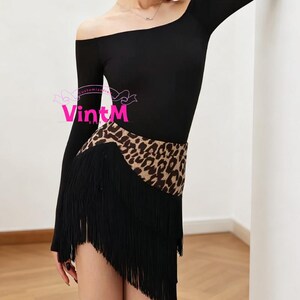 Puede incluir: Maillot de baile negro con escote asimétrico y mangas largas. La cintura presenta una banda con estampado de leopardo y una falda con flecos negros. El logo VintM es visible.