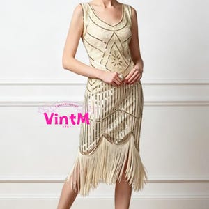 May include: A cream-colored, sleeveless flapper dress with a V-neckline. The dress features vertical sequin stripes and a geometric sequin design on the bodice. The skirt has a fringe hemline, creating a vintage look.