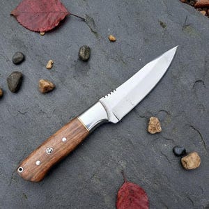 May include: A handcrafted hunting knife with a polished steel blade and a wooden handle. The handle is secured with rivets and features a brass bolster. The knife is shown on a dark gray stone surface.