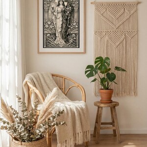May include: A framed black and white art print depicting a central figure holding a torch and flowers, surrounded by floral motifs and skulls. The print is displayed on a wall next to a cream-colored macrame wall hanging and a wooden stool.