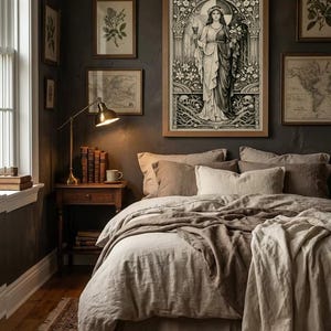 May include: A framed black and white art print depicting a central figure holding a torch and flowers, surrounded by ornate floral borders and skulls. The artwork is displayed on a dark wall above a bed with layered beige and brown bedding.