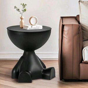 May include: A matte black side table with a rounded bowl-shaped top and a base featuring three distinct, curved feet. On the table sits a small beige vase with a green sprig, a circular wooden object, and a white book titled TICIN FOSDICE.