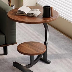 May include: A round side table with two tiers featuring dark wood grain surfaces and a black metal frame. The table has a curved support structure connecting the top and bottom circular shelves, resting on a flat, rectangular black metal base.