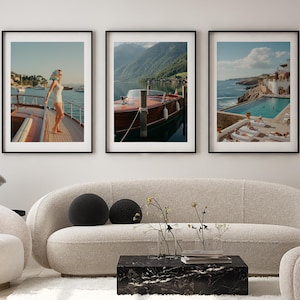 Könnte beinhalten: Drei gerahmte Kunstdrucke mit malerischen Küstenansichten. Einer zeigt eine Person auf einer Yacht, ein anderer ein Holzboot auf einem See und der dritte einen Pool am Meer. Die Drucke werden über einem cremefarbenen Sofa und einem schwarzen Marmor-Couchtisch ausgestellt.