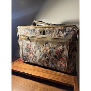 May include: A rectangular suitcase featuring a floral tapestry pattern in shades of red, blue, and beige. The luggage includes tan leather-like trim, a top handle, a front zippered pocket, and a small square logo emblem centered on the upper front.