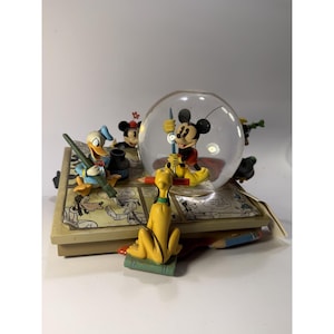 Könnte beinhalten: Ein dekoratives Disney-Schneekugel- und Figurenset mit Mickey Maus in einer Glaskugel sowie Donald Duck und Pluto auf einem Sockel, der wie eine Comic-Seite mit schwarz-weißen Illustrationen gestaltet ist.