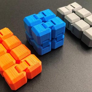 Könnte beinhalten: Drei farbenfrohe Zappelspielzeuge werden auf einer dunklen Oberfläche gezeigt. Ein orangefarbenes, ein blaues und ein graues Zappelspielzeug sind abgebildet. Das orangefarbene ist in einer geraden Linie, das blaue in Würfelform und das graue in einer geraden Linie.