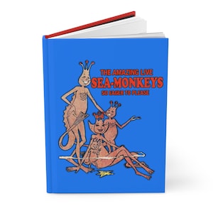 May include: A blue journal with the text "THE AMAZING LIVE SEA-MONKEYS SO EAGER TO PLEASE" and a vintage illustration of several Sea-Monkeys. The journal has a red spine and white pages.