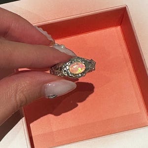 Pode incluir: Um anel de prata com uma pedra preciosa de opala oval e iridescente. O anel tem um design texturizado e orgânico. A opala exibe uma gama de cores, incluindo rosa, amarelo e laranja. O anel é segurado em uma caixa rosa.