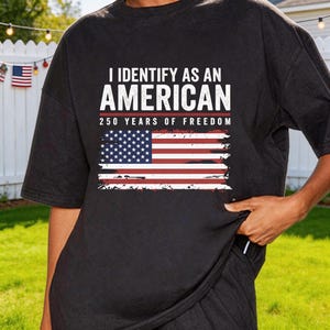Puede incluir: Camiseta negra con la frase "I IDENTIFY AS AN AMERICAN" en letras blancas sobre una bandera estadounidense desgastada. Debajo, el texto "250 YEARS OF FREEDOM". La camiseta se lleva al aire libre, en un jardín.