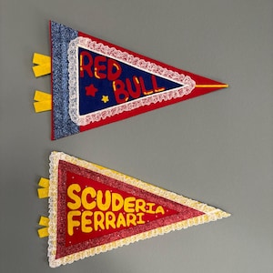 Può includere: Due gagliardetti triangolari. Il gagliardetto superiore è rosso con "RED BULL" in lettere bianche, un bordo blu e dettagli gialli. Il gagliardetto inferiore è rosso con "SCUDERIA FERRARI" in lettere gialle e dettagli gialli.