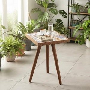 May include: A small, square side table with a mosaic tile top and three wooden legs. The table is holding a cup of coffee, a glass of water, and a book. The table is surrounded by various potted plants.