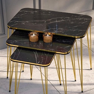 Puede incluir: Un juego de tres mesas nido con tapas negras con efecto mármol y patas de metal dorado. Las mesas tienen esquinas redondeadas y un borde dorado. Dos pequeñas velas están sobre la mesa del medio.
