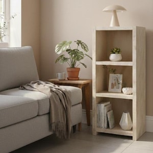 Puede incluir: Escena de salón en tonos neutros. Un sofá gris claro con una manta beige. Una mesa auxiliar de madera con una planta en maceta y una taza. Una estantería de madera clara con una lámpara de seta y una pequeña suculenta.