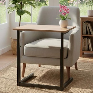 Puede incluir: Una pequeña mesa auxiliar ajustable con una tapa de madera clara y un marco de metal negro. La mesa está colocada junto a un sillón gris. Una pequeña planta en maceta con flores rosas está sobre la mesa. La mesa mide 61 cm de largo y 41 cm de ancho.