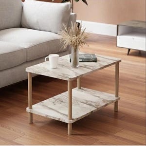 May include: A two-tiered coffee table with a marble-look top and a lower shelf. The table has light-colored wooden legs and supports. A white mug, a vase with dried plants, and a book are on the top surface.