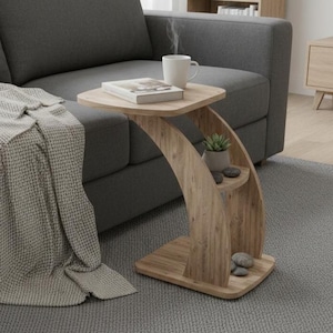 Pode incluir: Uma mesa lateral de madeira com um design curvo único. A mesa tem um tampo quadrado, uma prateleira pequena e uma base. Uma caneca branca com vapor, um livro e uma planta pequena estão sobre a mesa. A mesa está ao lado de um sofá cinza e de um cobertor cinza.
