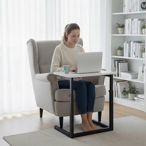 May include: A white and black side table with a laptop on top. The table is supported by a black metal frame. The person is sitting in a grey armchair in a well-lit room.