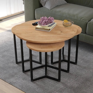 Puede incluir: Un juego de dos mesas de centro apilables. Las mesas tienen tapas redondas de madera marrón claro y patas de metal negro. La mesa más grande está encima de la más pequeña. Una alfombra gris está debajo.